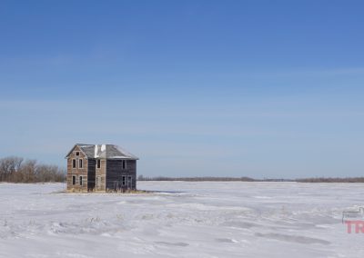 an abandoned, farm house with weathered wood siding sits in the middle of a flat, frozen farm field