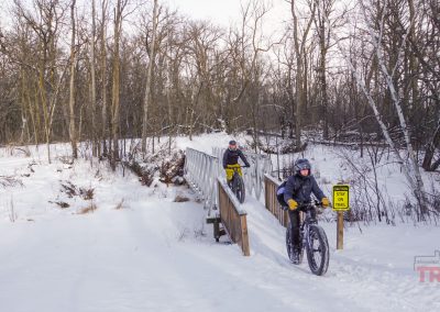 Two men on fatbikes cross a snow covered bridge on the edge of a forest