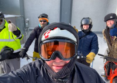 A man wearing snow goggles and a bike helmet takes a selfie with four other men holding fatbikes in the background