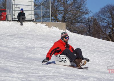 A man descends a snowy hill in a laundry basket mounted to a snowboard