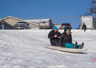 A man and a woman descend a snowy hill on a surfboard
