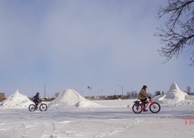 Two men riding fatbikes pass by snow sculptures on an open field