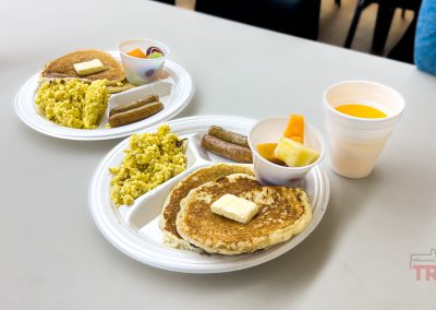 A closeup of two plates of sausage, eggs and pancakes and a fruit cup on a table