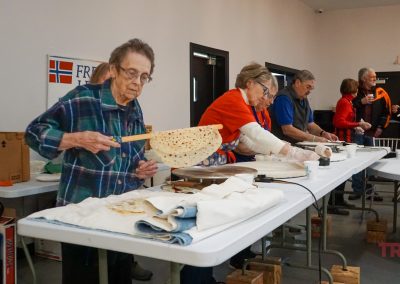 An elderly woman at the end of a lefse production line lifts a finished lefse off a griddle with a stick