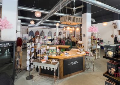 A boutique store interior featuring a wooden checkout counter with hanging lights above. Various clothing items and accessories are displayed on racks. Decorative pink cherry blossoms and a mounted deer head add a stylish touch.