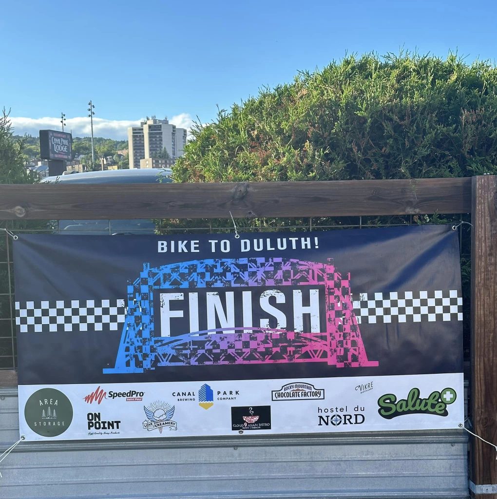 Bike to Duluth-Finish Line Banner reading Bike to Duluth! FINISH with a checkered flag design. Logos of various sponsors, including SpeedPro, Canal Park Brewing Company, and Salute, are displayed at the bottom. Trees and a few buildings are visible in the background.