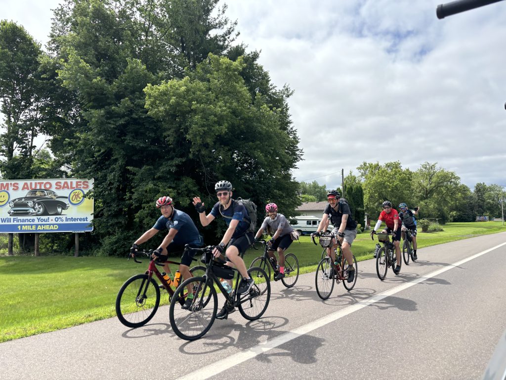 Bike to Duluth-Group Ride A group of six cyclists riding on a road surrounded by greenery. One cyclist in the middle waves at the camera. A sign for a car sales business is visible on the left, and the sky is partly cloudy.