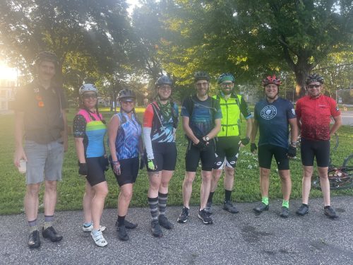 Bike to Duluth-Group A group of eight cyclists in colorful gear stand together in a park. They wear helmets and pose in front of a grassy area with trees. The sun is setting, casting a warm glow on the scene.