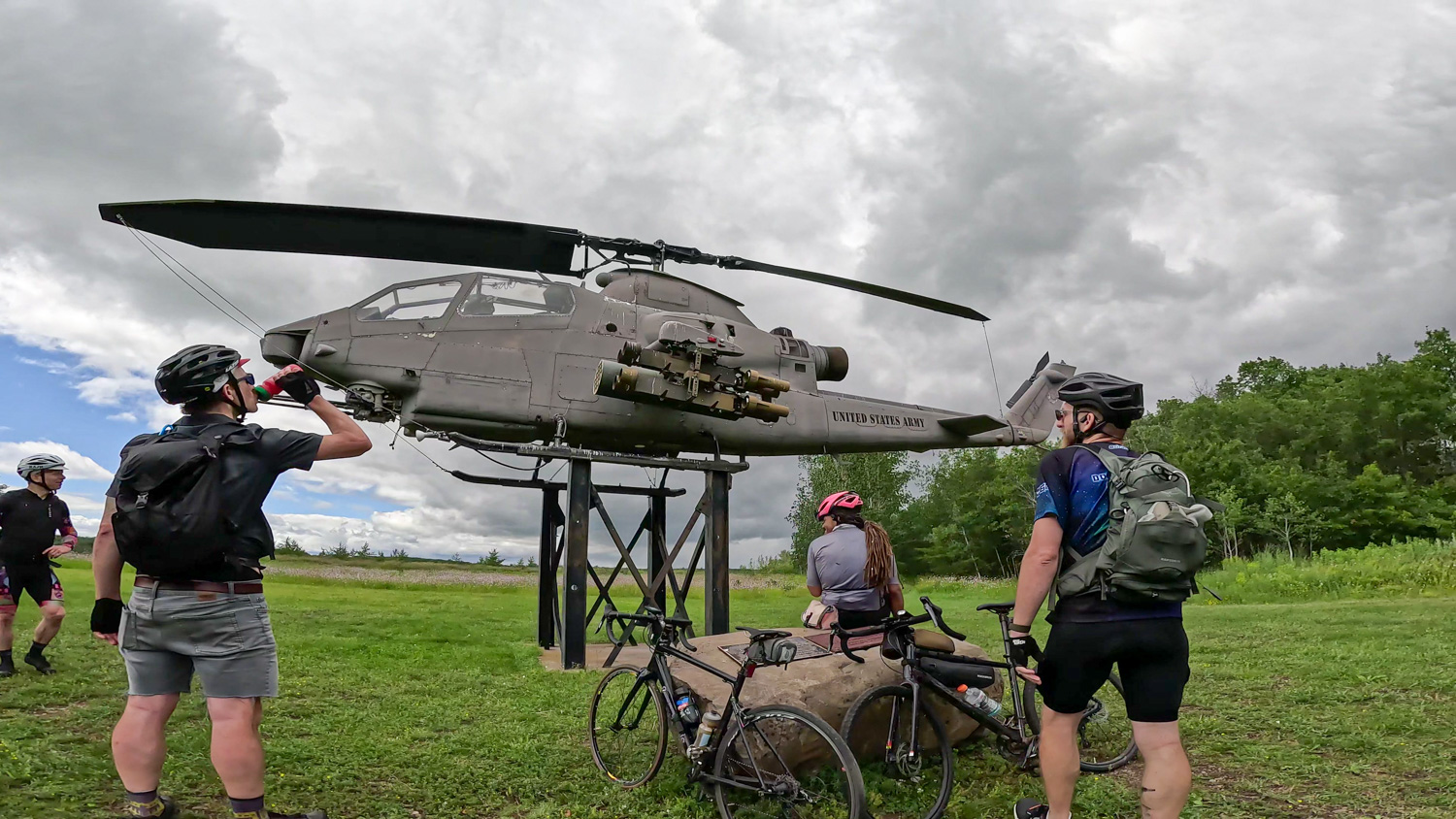 Bike to Duluth-Helicopter Cyclists pause on a grassy field beside a mounted military helicopter display. The sky is cloudy, and one cyclist drinks from a bottle. Their bikes are parked nearby, and there are trees in the background.