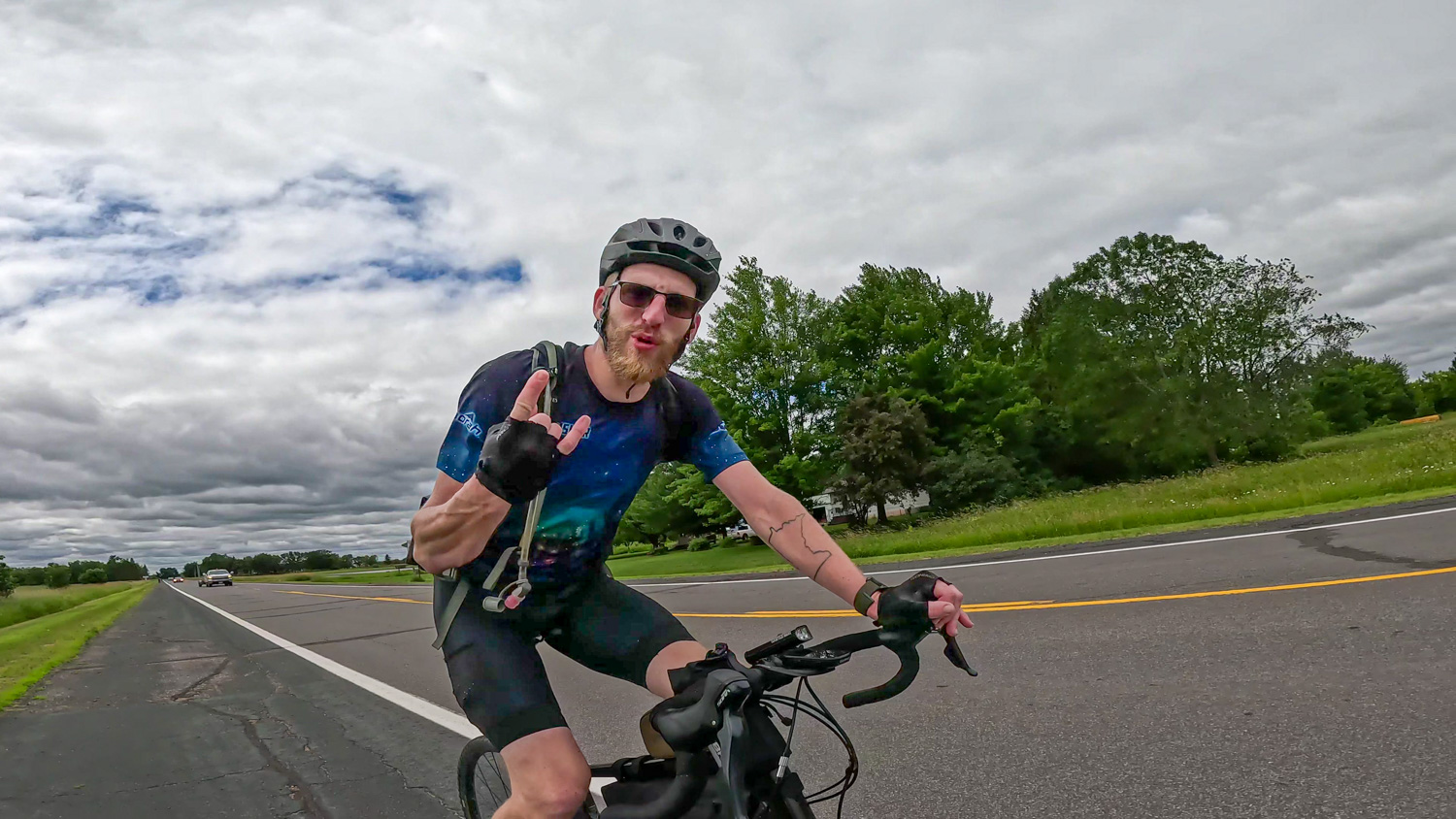 Bike to Duluth-Rock On A cyclist wearing a helmet, sunglasses, and cycling gear shows a hand gesture while riding on a road bordered by trees. The sky is cloudy, and a few cars are visible in the distance.