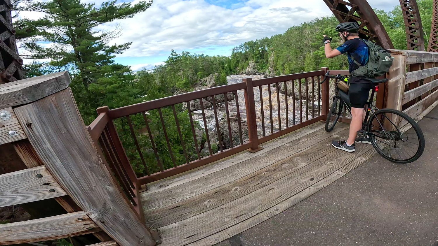 Bike to Duluth-St. Louis River Bridge Person with a bike and backpack photographs the St Louis River from a wooden bridge on the Willard Munger Trail. They wear a helmet and cycling gear. The scene is surrounded by lush green trees under a partly cloudy sky.
