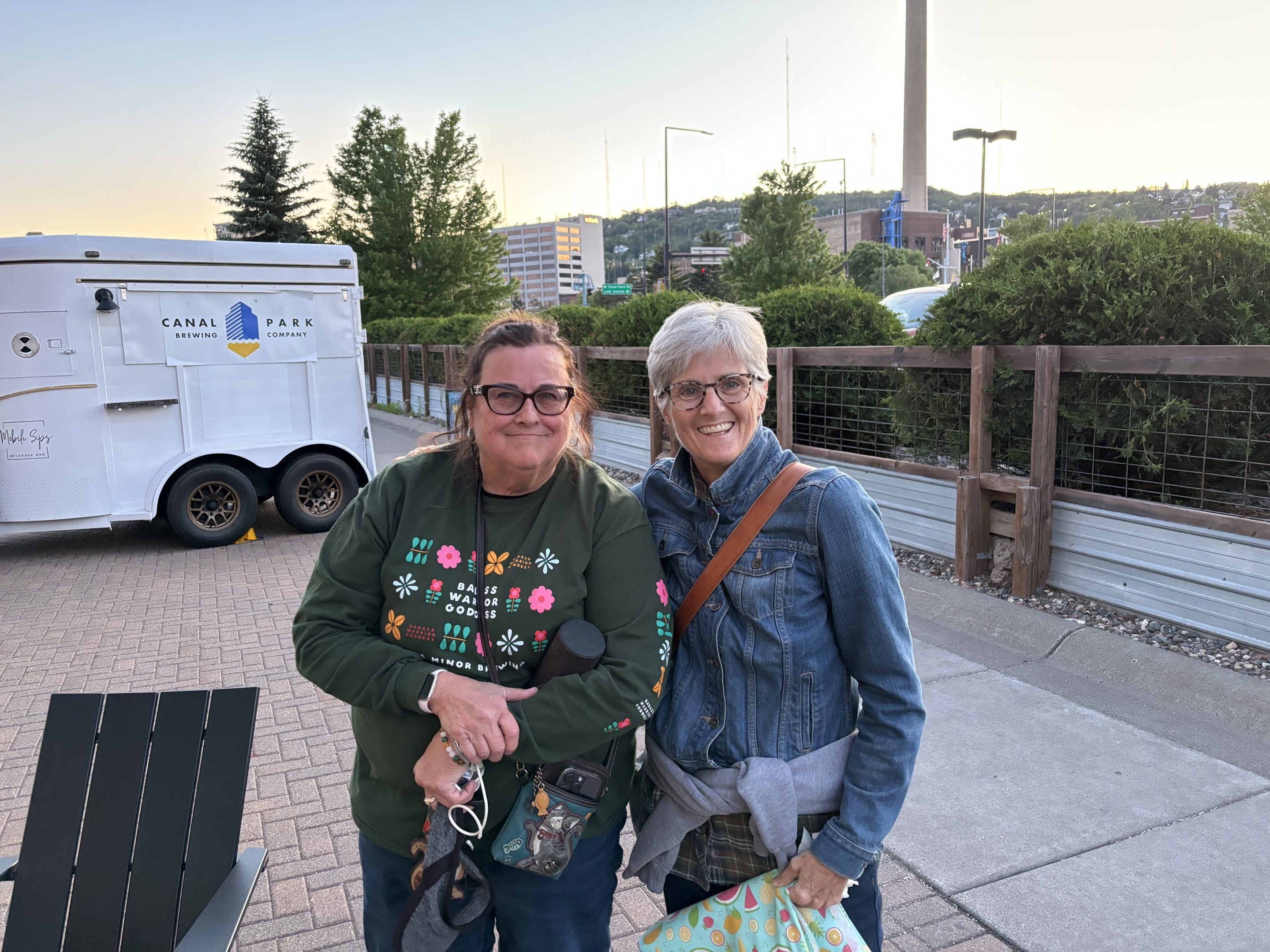 Bike to Duluth-Volunteers Two women smiling and standing together outdoors near a trailer with Canal Park Brewing Company on it. One wears sunglasses and a green sweatshirt; the other has glasses, a denim jacket, and carries a patterned bag. Trees and buildings are in the background.