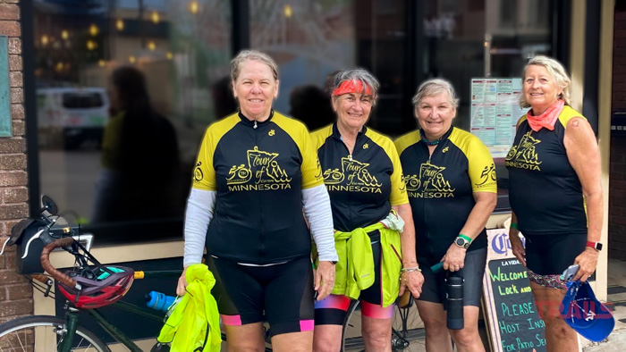 Tour of Minnesota Four women in matching Tour of Minnesota cycling jerseys and shorts stand beside a bicycle. They are outdoors, in front of a building with reflective windows. Two hold bright yellow clothing items, and one wears a red bandana.
