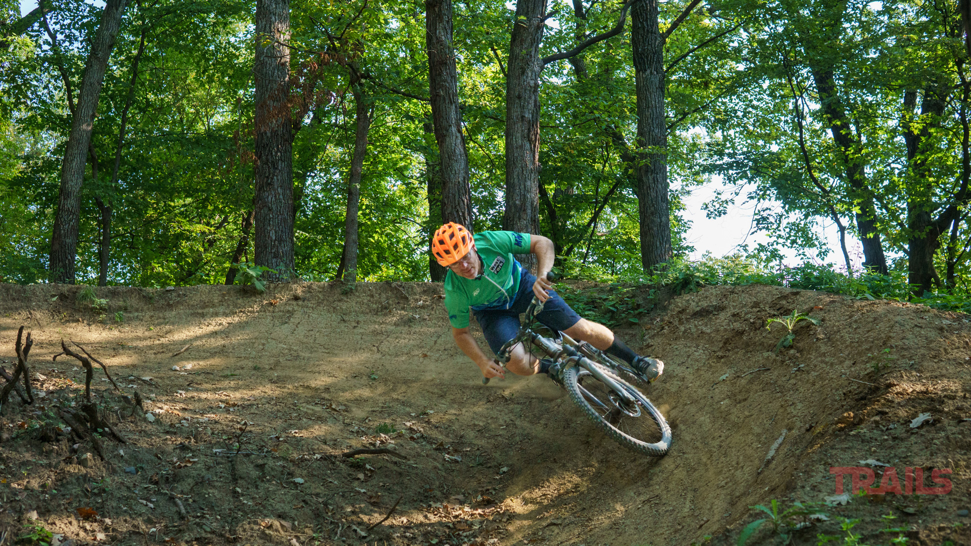 Belmont County Park A mountain biker wearing a green shirt and orange helmet leans into a turn on a dirt trail surrounded by tall, green trees in Belmont County Park’s lush forest.