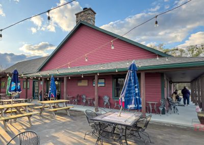 Outdoor dining area with wooden tables, metal chairs, and closed blue umbrellas in front of a red building at Grand View Valley Winery in Belview, MN. String lights hang overhead beneath a partly cloudy sky and beside a rustic stone chimney.