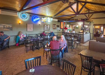 A cozy restaurant in Belview, MN with wooden ceilings, several tables and chairs, people dining and chatting, neon beer signs on the wall, and a small bar area. Enjoy the warm, casual atmosphere at Grand View Valley Winery.