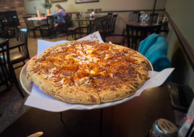 A close-up view of a large, cooked pizza on a metal stand at Grand View Valley Winery in Belview, MN, with tables, chairs, and a person sitting in the background. The pizza is topped with sauce and melted cheese.