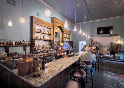 A bartender serves drinks to patrons at a marble bar in the stylish, warmly lit Rivendell Lounge in Belview, MN, with hanging lights, a large mirror, and shelves stocked with bottles. Guests chat and enjoy the relaxed atmosphere.