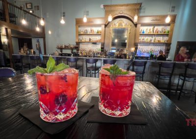 Two vibrant red cocktails garnished with mint sit on a dark wooden table at Rivendell Lounge in Belview, MN, with shelves of bottles, hanging lights, and patrons seated at the bar in the background.
