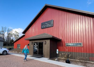 A person walks toward the entrance of the Rivendell lounge atGrand View Valley Winery in Belview, MN. A large red and brown building with a peaked roof on a clear day. A white car is parked nearby, and a sign with a logo is above the entrance.
