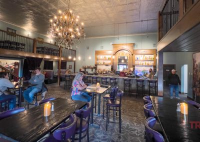 Spacious bar with high ceilings, chandelier, and balcony seating. Several people sit at tables and the bar. Behind the bar: lights and shelves of bottles. Decor includes candles on tables and a mural at the Rivendell Lounge at Grand View Valley Winery in Belview, MN.