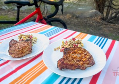Two plates with grilled pork chops and pasta salad sit on a colorful striped tablecloth outdoors, beside a red bicycle and near a riverbank with trees in the background.