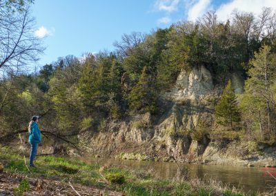 A person in a teal jacket and jeans stands on a grassy riverbank in Ramsey Park, Redwood Falls, Minnesota, gazing across the Redwood River at steep, tree-covered cliffs under a partly cloudy sky.