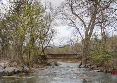 A wooden footbridge spans flowing Ramsey Creek surrounded by rocks and leafing trees in early spring at Ramsey Park/Cansayapi in Redwood Falls, Minnesota. A person in blue is walking across the bridge under a cloudy sky.