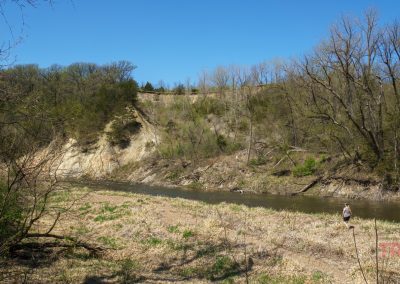 A river flows beside a steep, sandy bluff covered with sparse trees and shrubs under a clear blue sky in Ramsey Park/Cansayapi near Redwood Falls, Minnesota. Leafless and budding trees line the riverbank, with grassy vegetation covering the ground.