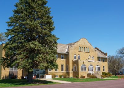 The Redwood County Historical Museum in Redwood Falls, MN, is a large yellow brick building with arched windows and an ornate roofline, tucked behind a tall evergreen tree and surrounded by green grass and bare trees beneath a clear blue sky.