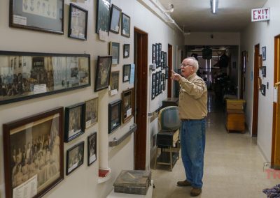 A man with grey hair stands in a hallway at the Redwood County Historical Museum in Redwood Falls, MN, looking at framed photographs displayed on the wall. The hallway is lined with doors and various items, and an exit sign is visible on the ceiling.