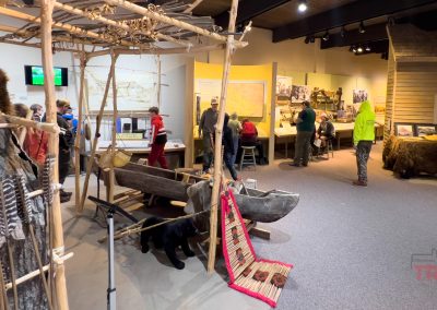 Visitors explore Lower Sioux Agency exhibits in Morton, MN, featuring historical artifacts like canoes, tools, and maps. Several people read displays and interact with exhibits in a well-lit room with rustic decor and informational panels.