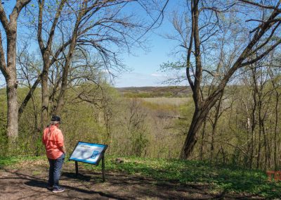 A person in an orange jacket reads an informational sign at the Lower Sioux Agency Historic Site near Morton, MN, standing at a scenic overlook with rolling hills and a forested landscape beneath a clear blue sky.