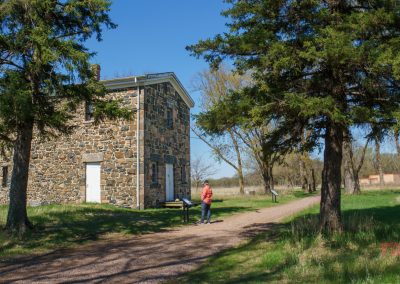A person stands on a gravel path near the historic Lower Sioux Agency Historic Site in Morton, MN, surrounded by grass and tall trees on a sunny day, reading a plaque by a stone building.