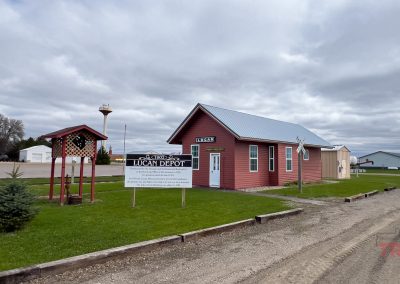 A small red historic depot building labeled “Lucan Depot” stands near a sign and covered bench, surrounded by green grass under a cloudy MN sky in rural Lucan, home of the Lucan Historical Society.