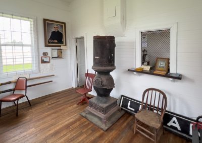 A small museum room in Lucan, MN, features wooden floors, old chairs, a wood stove, framed photos, rifles on the wall, historical papers, and a window letting in natural light—preserved by the Lucan Historical Society.