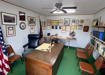 A small office in Lucan, MN with an American flag, a wooden desk, chairs, shelves of books, and walls covered in framed photos and plaques from the Lucan Historical Society. A ceiling fan hangs above and a typewriter sits on the desk.