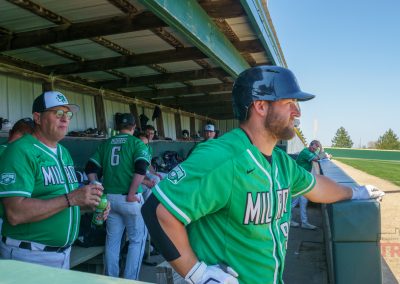 A baseball player in a green jersey stands by the dugout fence at The Irish Yard baseball stadium in Milroy, MN, with teammates and coaches in matching uniforms inside the dugout on a sunny day.