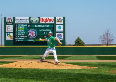 A baseball pitcher in a green and white uniform winds up on the mound at The Irish Yard baseball stadium in Milroy, MN, as an infielder stands ready. A large scoreboard in the background displays team lineups and game information on a sunny day.