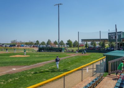 A baseball game is in progress on a sunny day at The Irish Yard baseball stadium in Milroy, MN. Players are on the field as fans watch from bleachers, with a scoreboard, team dugouts, and parked cars visible in the background.