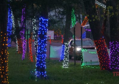 Trees lining a road in Redwood Falls are wrapped in colorful holiday lights—blue, orange, purple, green, and red—while a car drives through the scene at the NightFalls event at Ramsey Park/Cansayapi in Redwood Falls, Minnesota.
