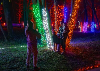 A person snaps a photo of a family standing among trees wrapped in colorful string lights at the NightFalls event in Ramsey Park/Cansayapi, Redwood Falls, Minnesota. A parked car is visible in the background under the night sky.