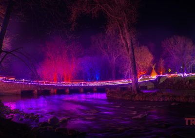A nighttime scene at Ramsey Park/Cansayapi shows a river flowing under a small bridge decorated with colorful lights, casting purple, pink, and red glows on the water and leafless trees in Redwood Falls, Minnesota during the NightFalls event.