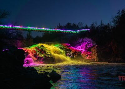 A waterfall at night in Ramsey Park/Cansayapi, Redwood Falls, Minnesota, is illuminated by colorful lights with a bridge above also decorated brightly during the NightFalls event.
