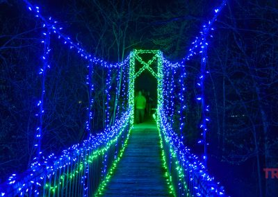 A wooden suspension bridge at night, glowing with blue and green string lights during the NightFalls event in Redwood Falls, Minnesota. Two people stand at the far end, framed by leafless trees.