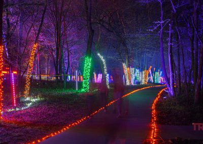 A forest path in Ramsey Park/Cansayapi, Minnesota, is lined with trees wrapped in vibrant string lights at night. The glowing trail edges light the way as faint, ghostly figures wander through Redwood Falls’ enchanting woods during the Nightfalls event.