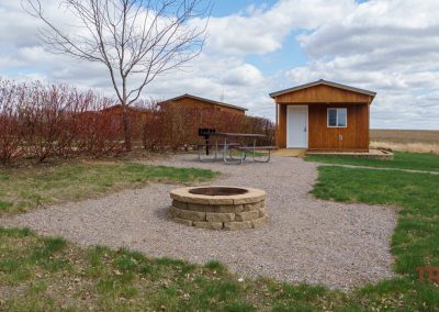 Several small wooden cabins sit near a picnic table and grill at Plum Creek Park in Walnut Grove, MN, with a circular stone fire pit in the foreground. Leafless trees and shrubbery surround the area; open field and cloudy sky in the background.