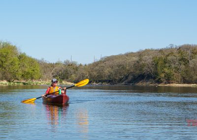 A person wearing a life jacket paddles a red kayak on the calm waters of Lake Redwood, surrounded by green trees and under a clear blue sky near Redwood Falls, MN.