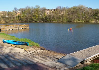 A calm river at Lake Redwood, MN, with a kayak launch ramp, a blue kayak on shore, a person kayaking on the water. A wooden dock and trees with spring foliage stand nearby under a clear blue sky in Redwood Falls.