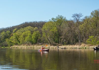 A person paddles a red kayak on calm Lake Redwood near Redwood Falls, MN, bordered by green trees and grassy banks under a clear blue sky.